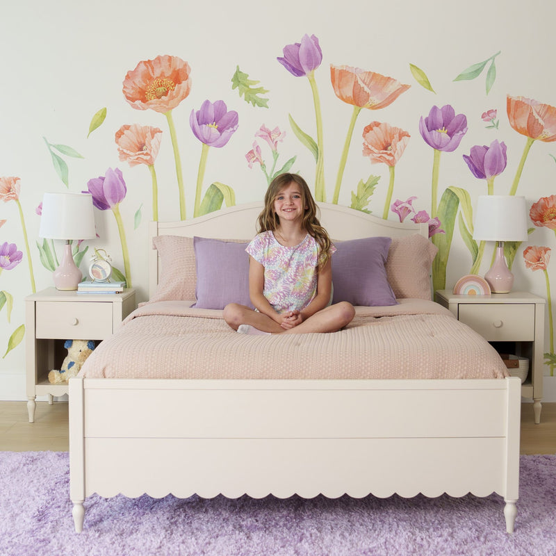 Young girl on pastel pink bed in girls' bedroom with watercolor floral wall decals, purple pillows and bedside lamps, kids bedroom decor