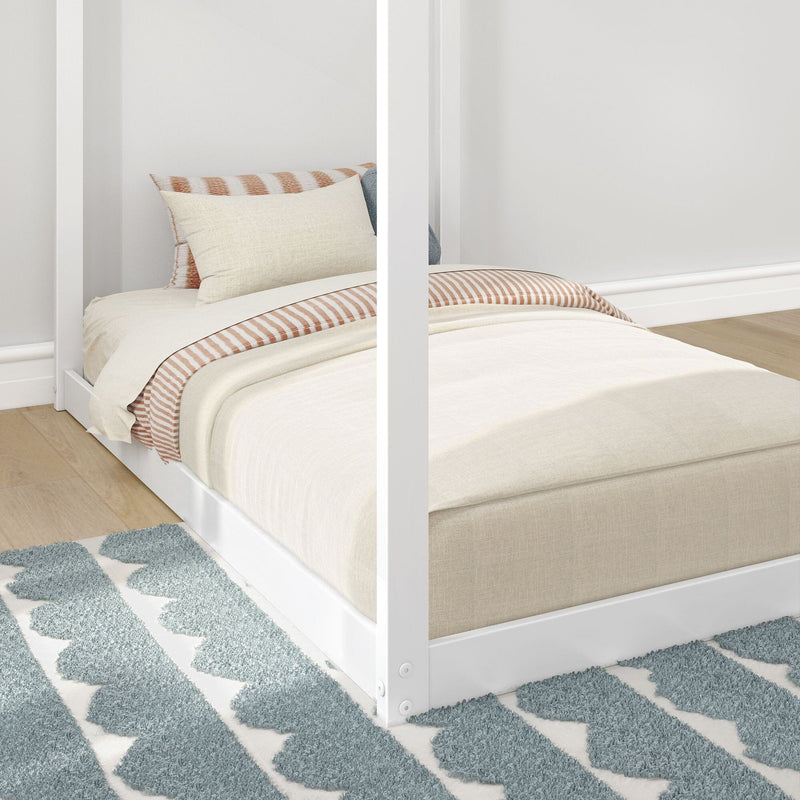 White four-poster bed with beige duvet, striped tan-and-white pillows, light wood floor and blue patterned rug in minimalist bedroom