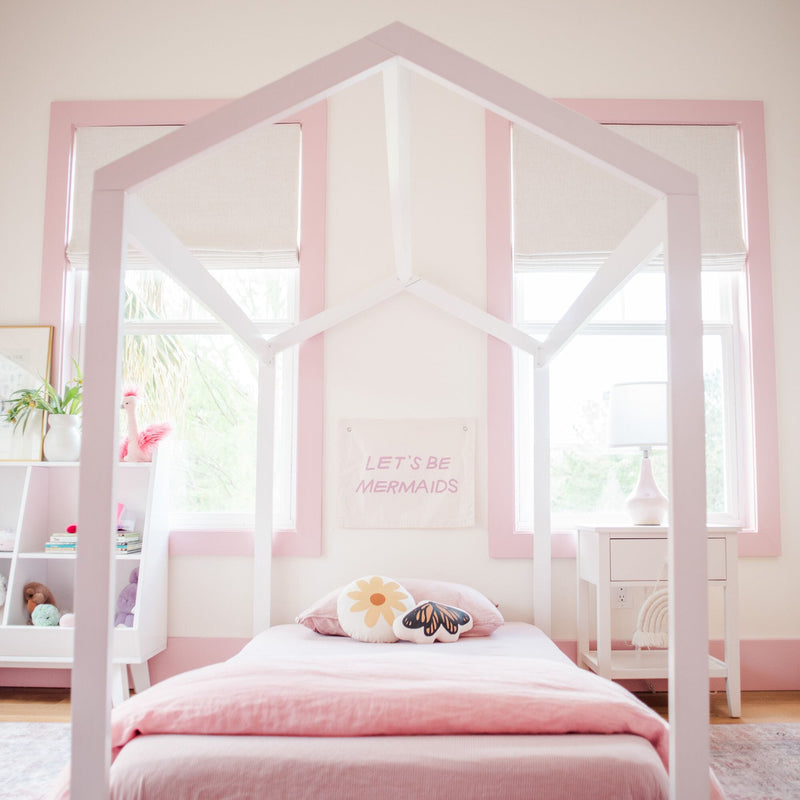 Pastel pink girl's bedroom with house-shaped white bed frame, soft pink bedding, daisy and butterfly pillows, "Let's Be Mermaids" wall art and white nightstand