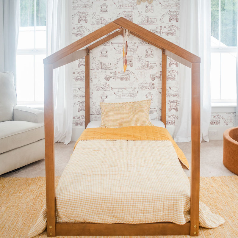 Wooden house-frame children's bed with yellow gingham bedding and construction truck wallpaper in a bright kids' nursery