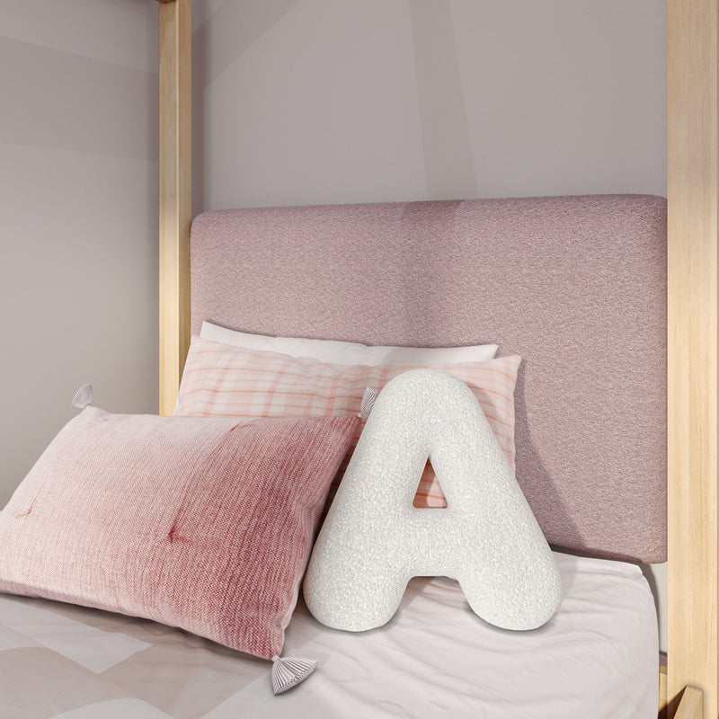 Blush pink upholstered headboard close-up with natural wood bedframe posts, soft pink gingham and textured mauve pillows, and a white decorative letter A cushion on pastel bedding