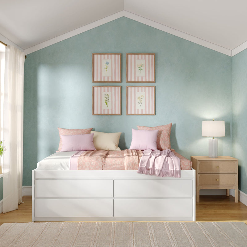 Pastel bedroom with mint-green accent wall and four framed botanical prints, white storage bed with pink floral bedding and pillows, wooden nightstand with table lamp and sheer curtains