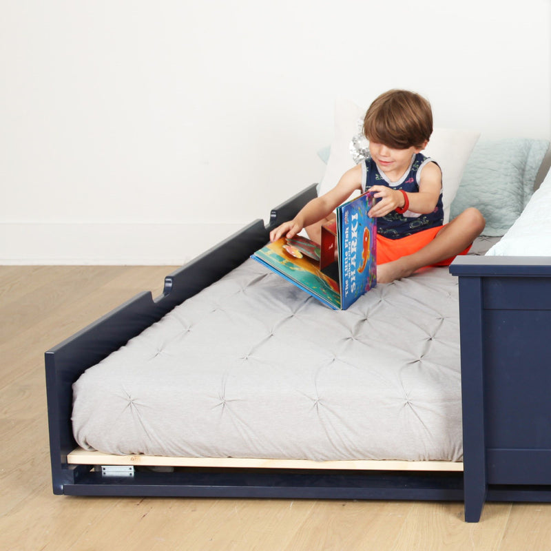 Child reading colorful children's books on a navy wooden toddler bed with tufted gray mattress in a bright kids' bedroom