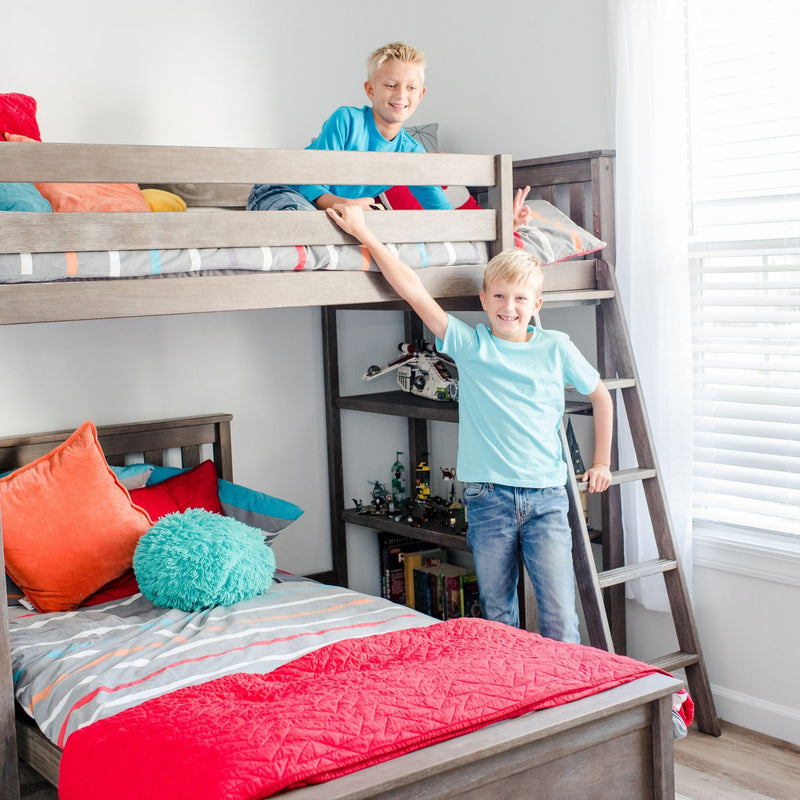 Two smiling boys on a gray wooden bunk bed in a bright kids' bedroom with colorful bedding, ladder and playful decor