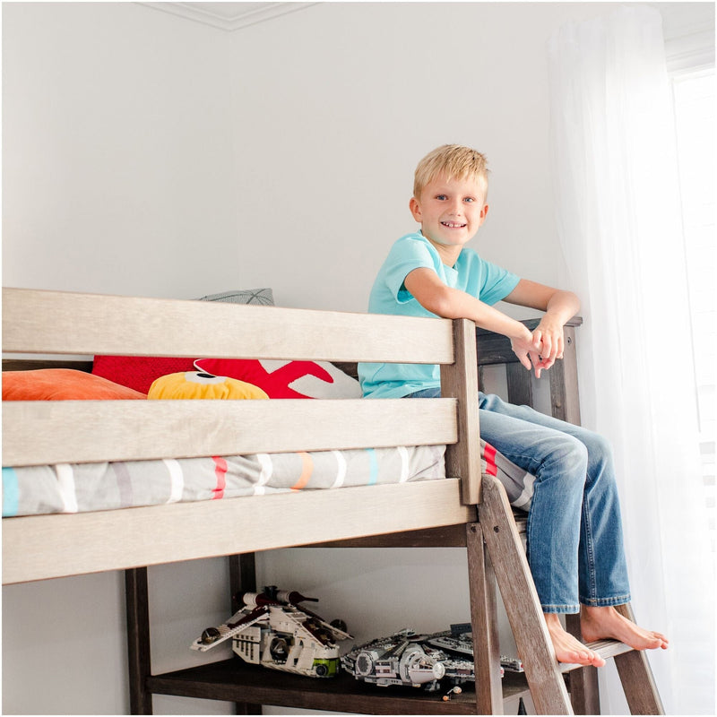 Smiling boy on a wooden bunk bed in a bright kids' bedroom with ladder, colorful pillows and model spaceship toys underneath