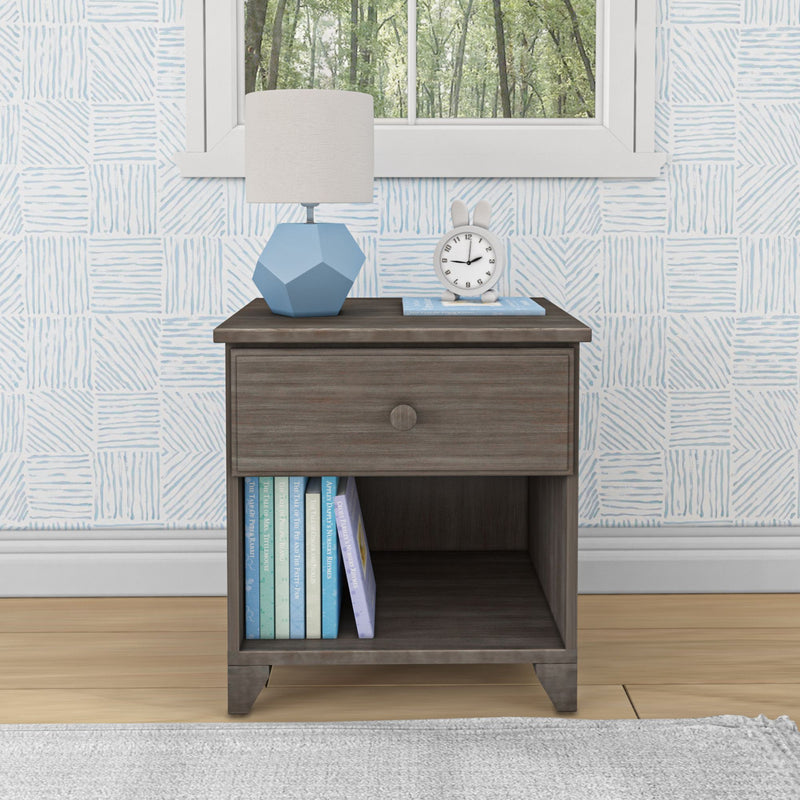 Nursery nightstand with blue geometric table lamp, white bunny-ear alarm clock, pastel children's books on the lower shelf, light-blue patterned wallpaper and a window overlooking a green forest