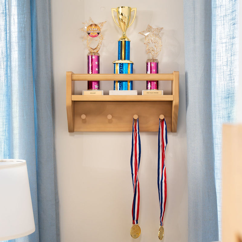 Gold cup trophy and two pink-and-blue star cheerleader trophies on a wooden wall shelf with two red-white-blue hanging medals and light blue curtains