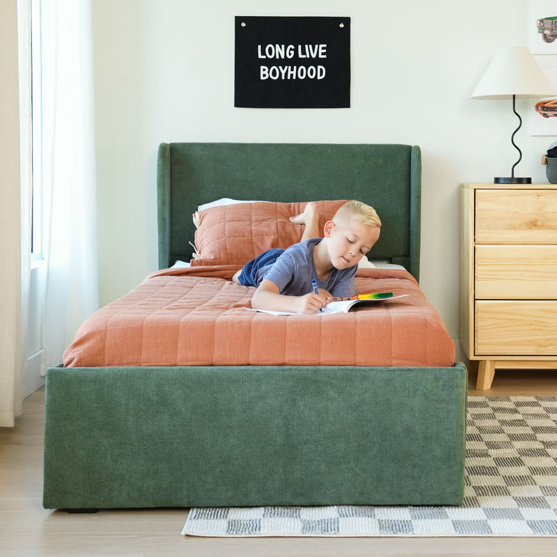 Young boy coloring on a bed with a green upholstered headboard and coral quilt beneath a "LONG LIVE BOYHOOD" wall banner in a bright modern bedroom