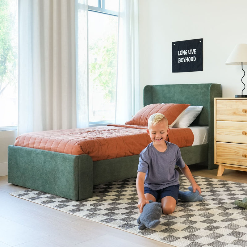 Child playing with stuffed toy on a checkered rug in a bright modern kids' bedroom with green upholstered bed, orange quilt and "Long Live Boyhood" wall sign