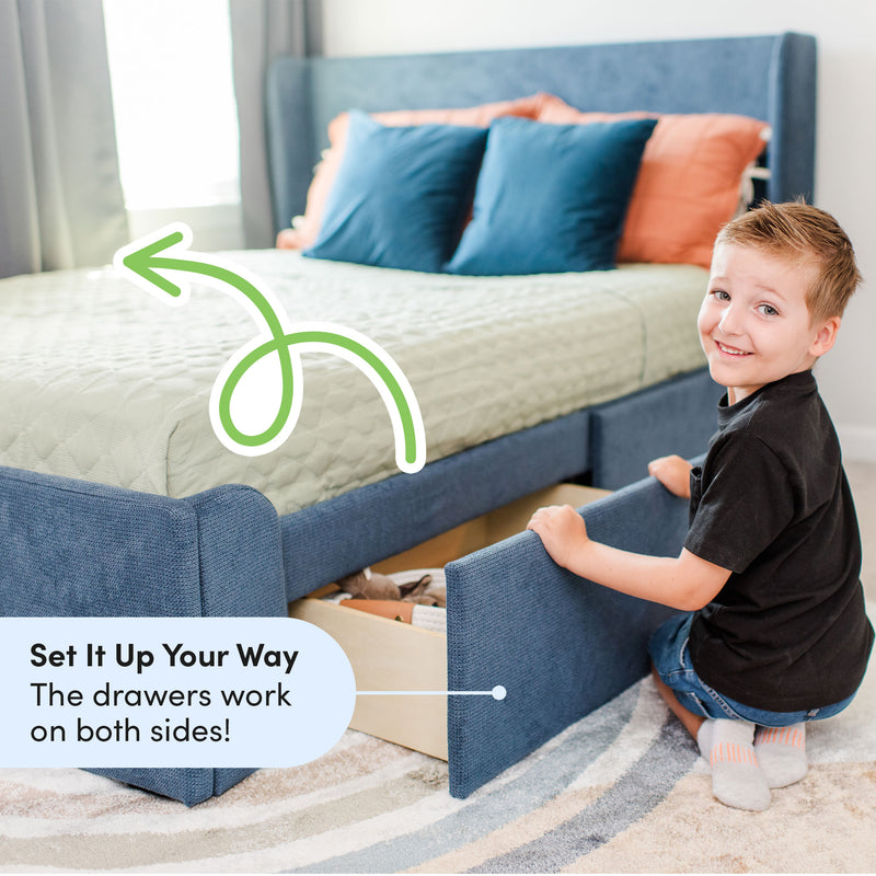 Smiling young boy opening an under-bed storage drawer on a blue upholstered storage bed with teal pillows — modular kids' bedroom solution with drawers on both sides