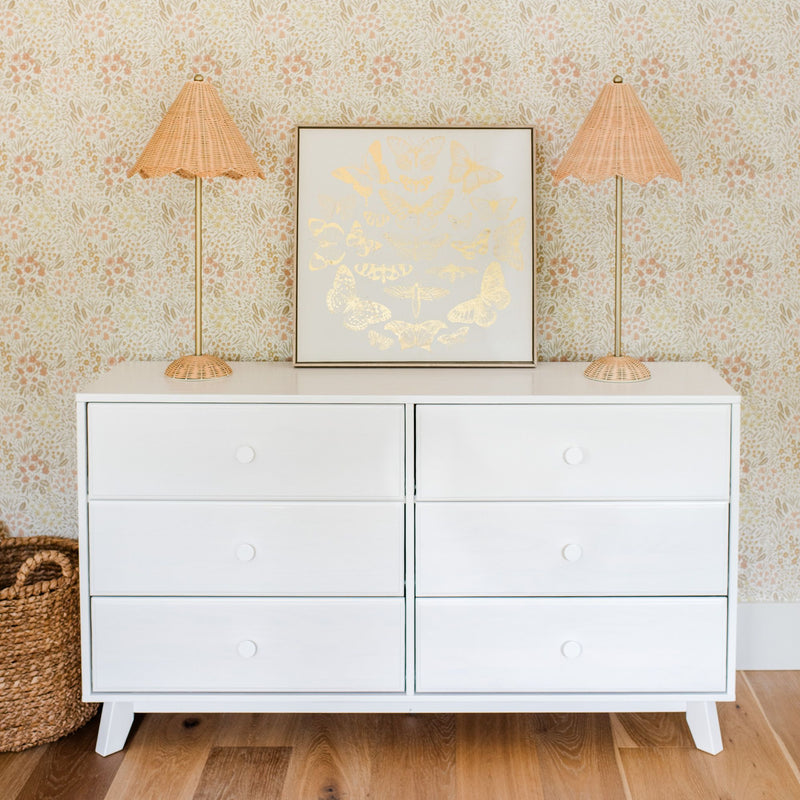 White six-drawer dresser styled with two wicker rattan lamps and framed gold butterfly art against pastel floral wallpaper — boho nursery décor
