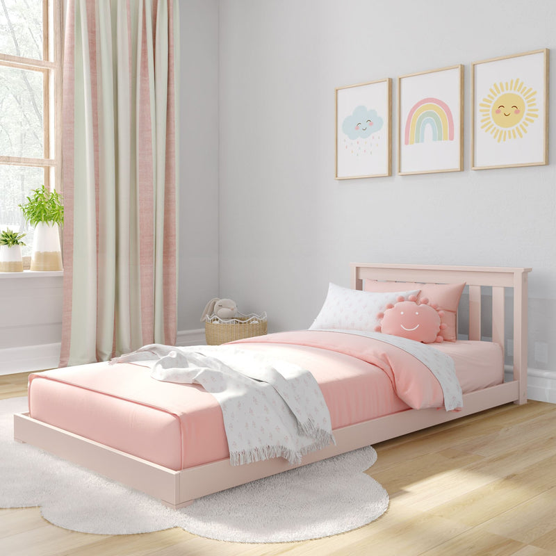 Pastel pink toddler bed and bedding in cozy nursery with smiling sun, rainbow and cloud wall art, blush curtains and potted plants