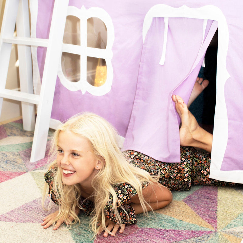 Smiling child with long blonde hair crawling out of a purple indoor play tent on a colorful geometric rug in a children's playroom