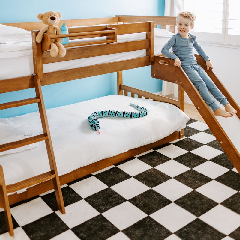 Smiling child on a wooden bunk bed with slide in a bright kids' bedroom featuring a checkered rug, plush toys, and blue walls