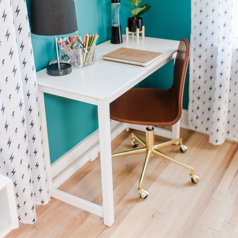 White home office desk with brown swivel chair, black lamp, clear container of colored pencils, spiral notebook and lightning-bolt curtains against a teal wall