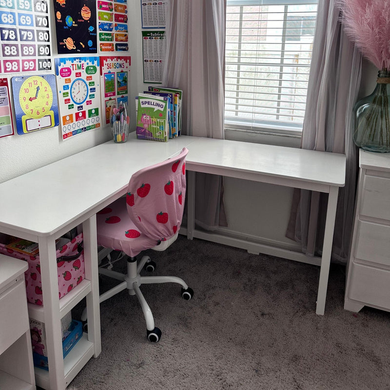 Bright kids' homeschool study corner with white L-shaped desk, pink strawberry swivel chair, educational posters (numbers, clock, planets, seasons), spelling workbook and markers by a window with blinds