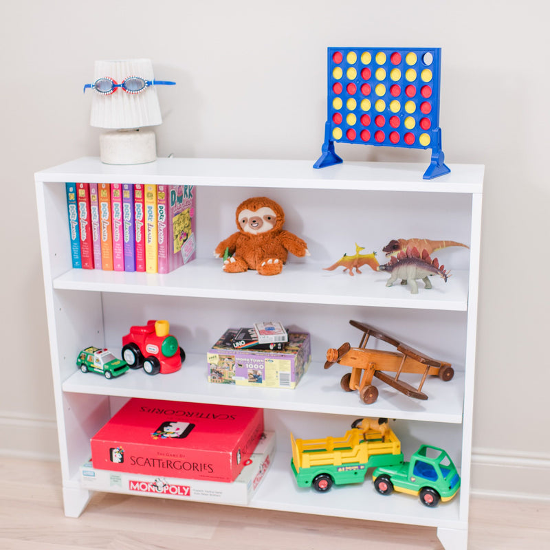 White playroom bookshelf filled with children's toys and games: Connect Four, colorful board games, stuffed sloth, toy dinosaurs, wooden airplane and toy trucks.