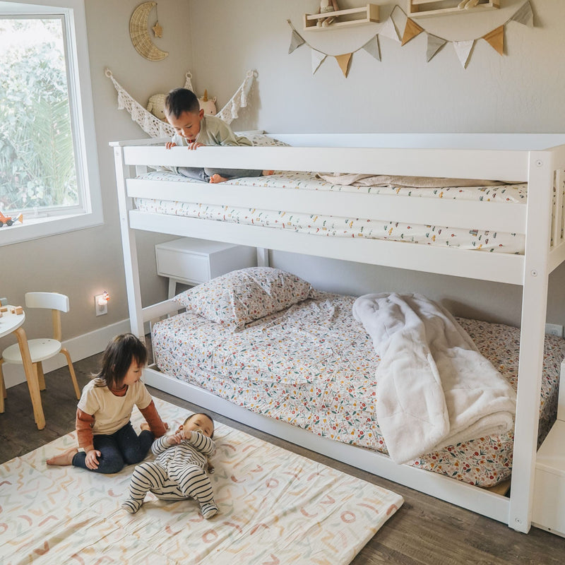 Children playing on a play mat in a neutral modern kids' bedroom with a white bunk bed, floral bedding, bunting wall decor, and natural window light