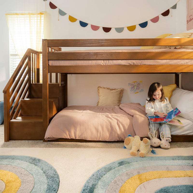 Wooden loft bunk bed for kids with built-in storage stairs, pastel pink bedding, rainbow rug, bunting wall decor and plush toys with a child reading in a cozy children’s bedroom reading nook