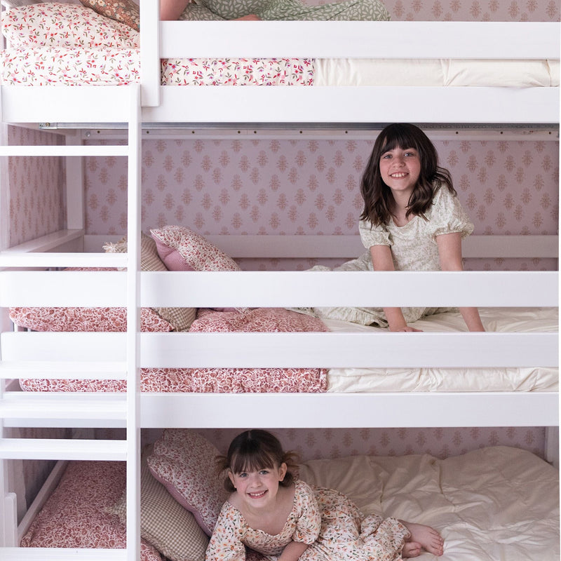 Kids bunk beds: two smiling girls lounging on white wooden bunk beds with floral bedding, pink wallpaper and cozy children's room decor