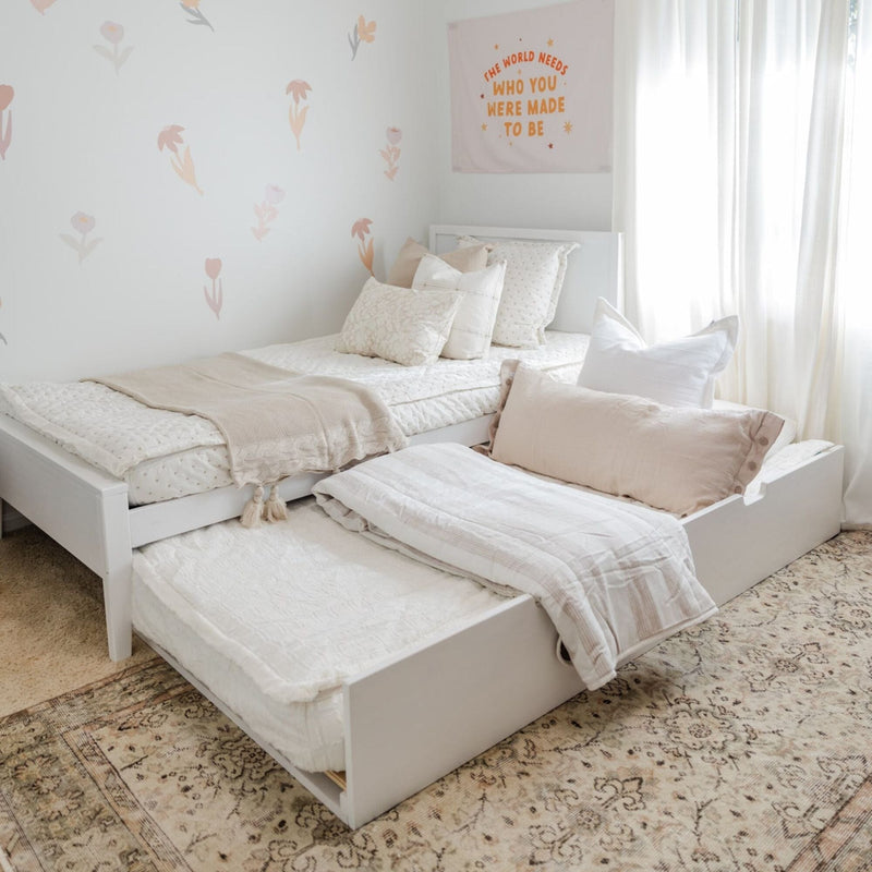 Neutral children's bedroom with white trundle beds, layered cream bedding, beige knit throw, soft pink pillows, floral wall decals and sheer curtains
