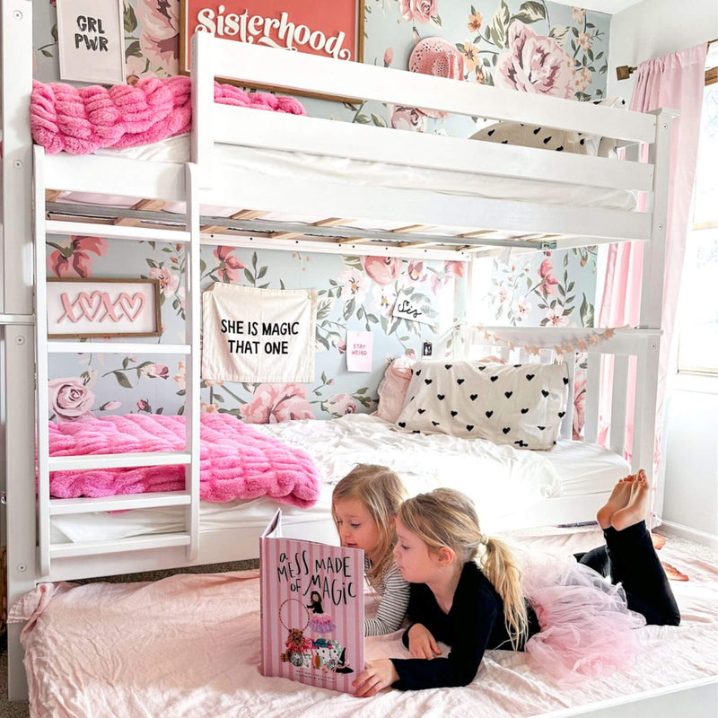 Girls bunk bed in pink floral bedroom with white loft frame, cozy pink blanket, heart-print pillows and two children reading, kids bedroom decor inspiration