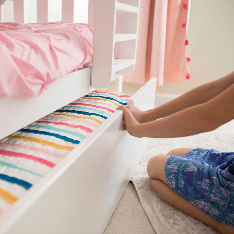 Child pulling out a trundle bed drawer with a colorful striped mattress beneath a white bunk bed with pink bedding