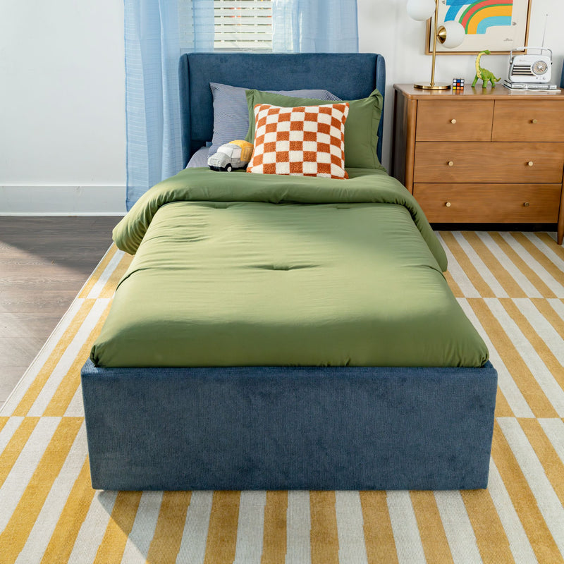 Kids bedroom with green bedding on a blue upholstered twin bed, orange checkered throw pillow, wooden dresser with lamp, retro radio, dinosaur toy and yellow striped rug