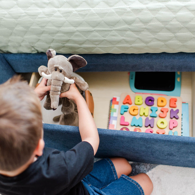 Child holding a plush elephant toy beside a colorful wooden alphabet puzzle under a quilted mattress in a kids playroom