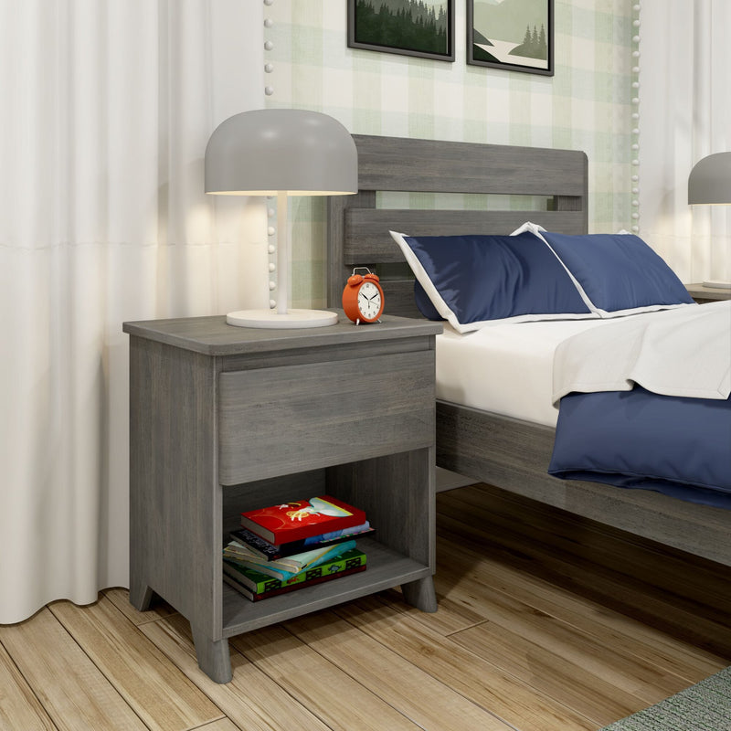 Gray wooden nightstand with lamp and red alarm clock beside bed with navy pillows, white duvet and colorful books on hardwood floor