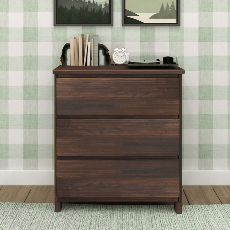 Dark walnut three-drawer dresser with books, geode bookends, alarm clock and record player against green plaid wall with framed nature prints