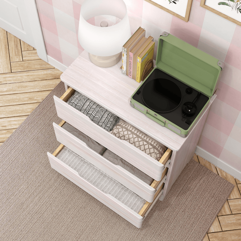 White bedroom dresser with open drawers, folded linens, green portable record player, lamp and books on pink checkered wall