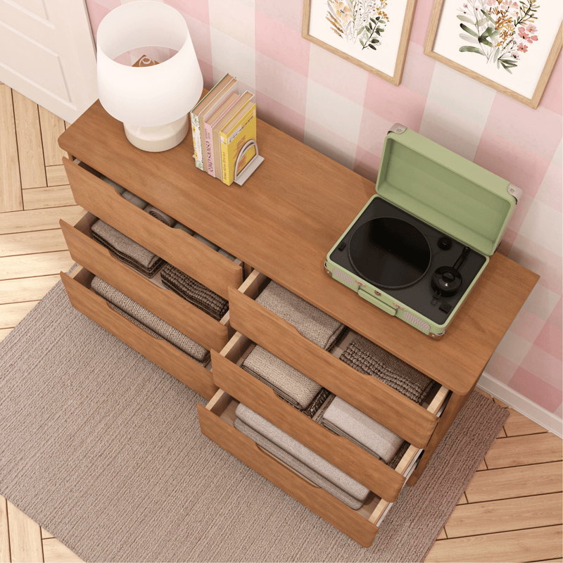 Top-down view of a wooden dresser with open drawers filled with neatly folded blankets, a pastel green portable record player, lamp and books beside floral framed art on pink checkered wallpaper in a cozy bedroom interior