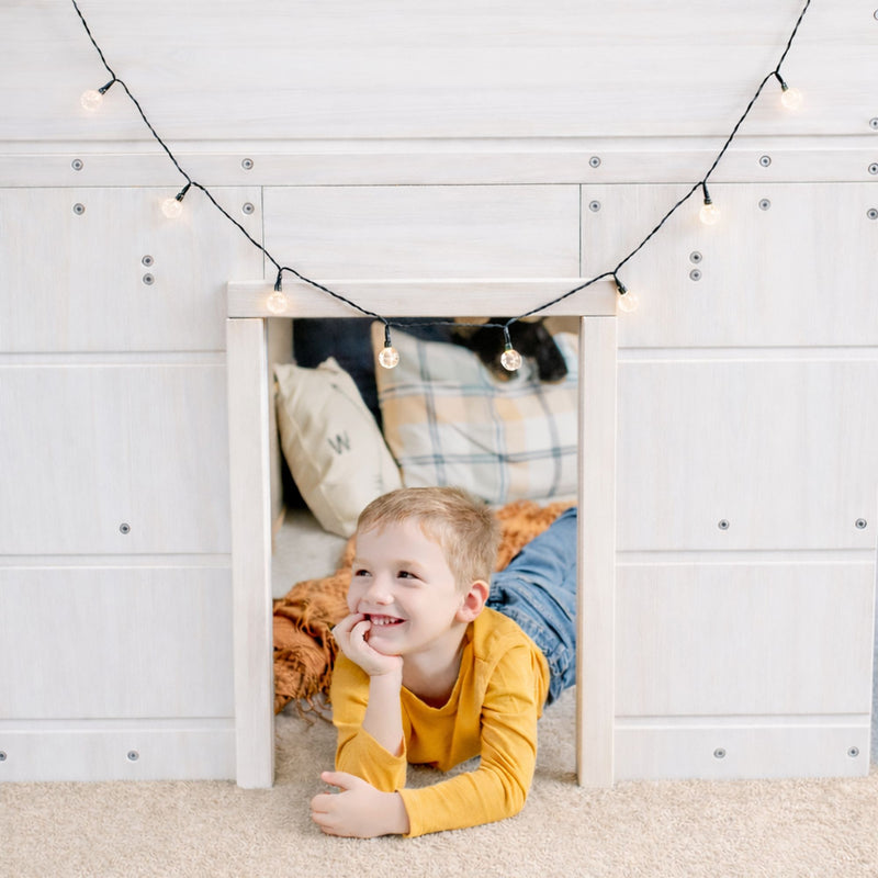 Smiling young boy in yellow shirt lounging inside a white wooden playhouse with cozy pillows and hanging string lights