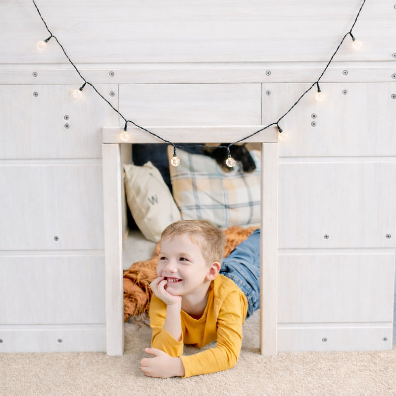 Smiling child in cozy indoor playhouse with hanging string lights, white wood paneling and plaid pillows, kids room decor
