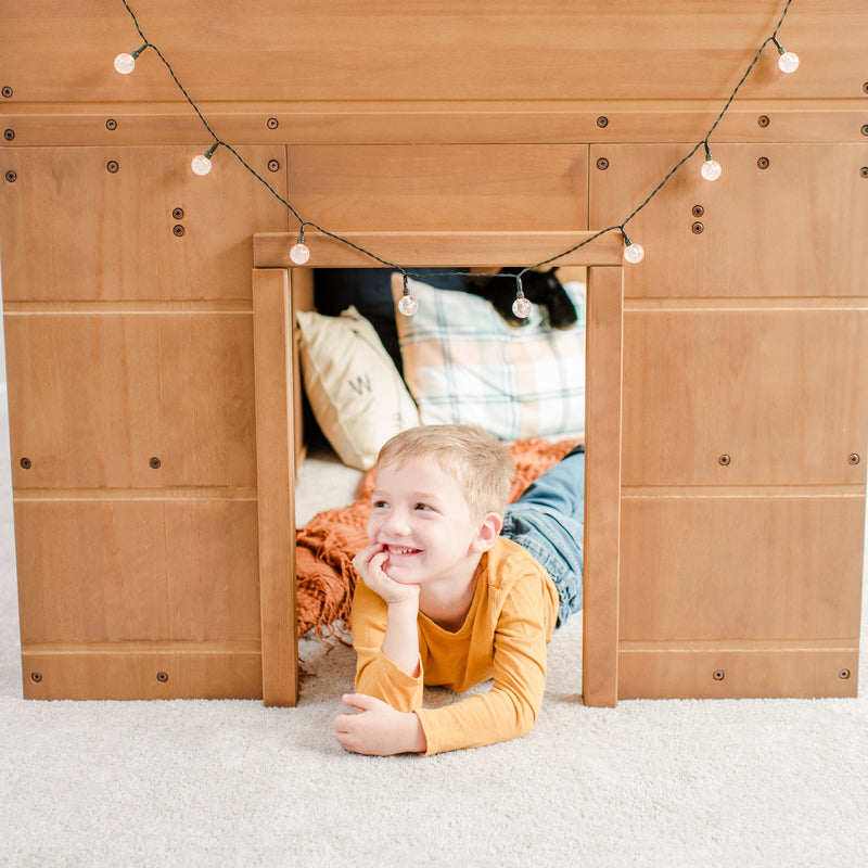 Smiling young boy lying in a wooden playhouse doorway with string lights and cozy pillows in a kids' indoor playroom