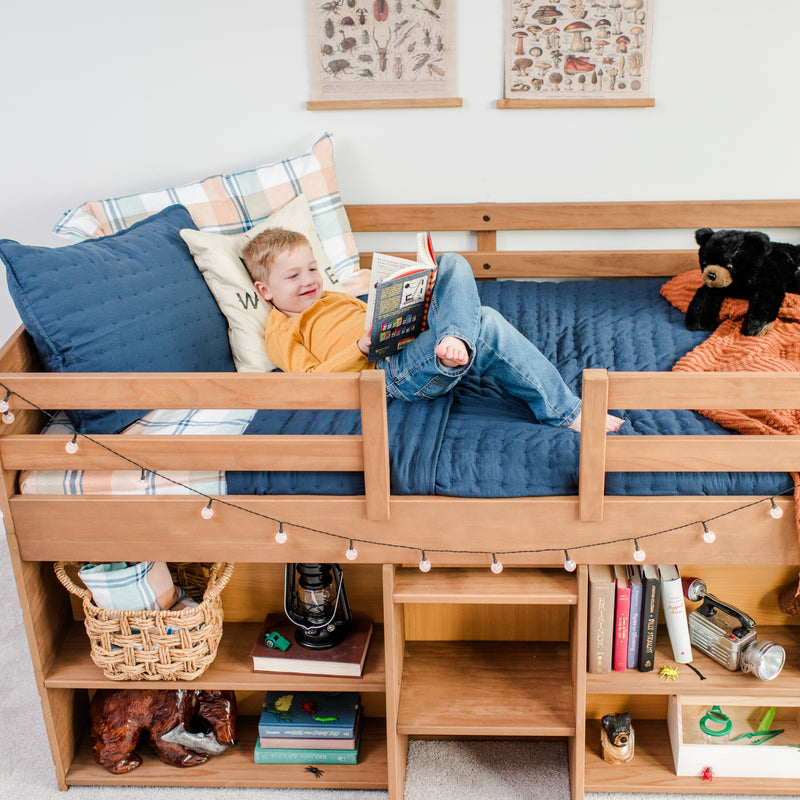 Child reading a book on a wooden loft bed in a cozy kids' bedroom with blue quilt, plaid pillows, teddy bear, string lights and built-in shelves with books and toys