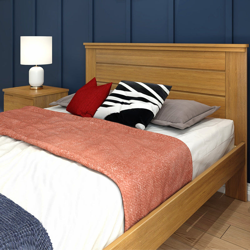 Close-up of a modern bedroom featuring an oak wooden bed frame and headboard, coral throw blanket, red and black-striped decorative pillows, white bedside lamp, and navy paneled wall