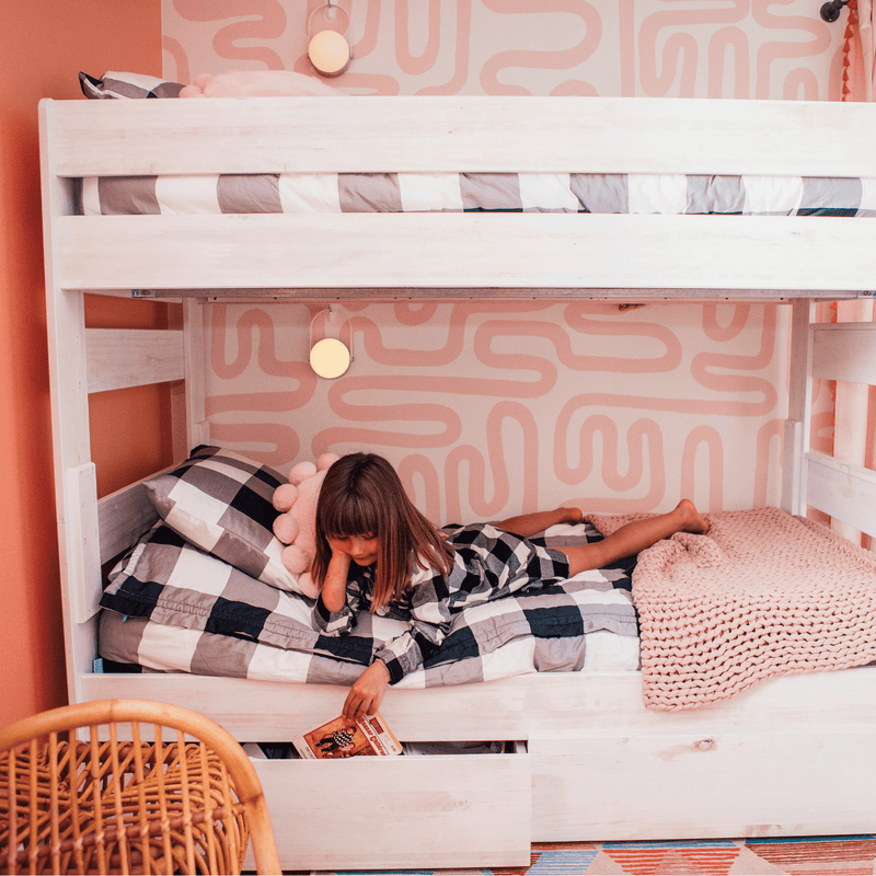 Child reading on lower bunk bed in pink-patterned kids bedroom, plaid bedding, knitted blanket and cozy nursery decor