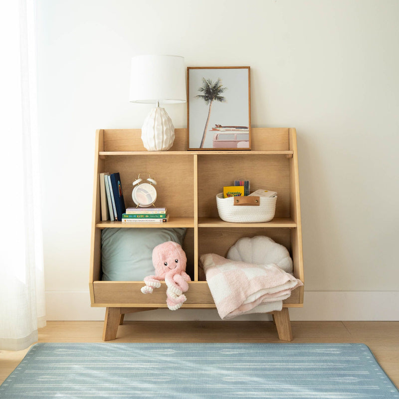 Wooden nursery cubby shelf with white ceramic table lamp, framed palm print, children's books and clock, woven basket, pink plush octopus and pastel blanket on blue rug