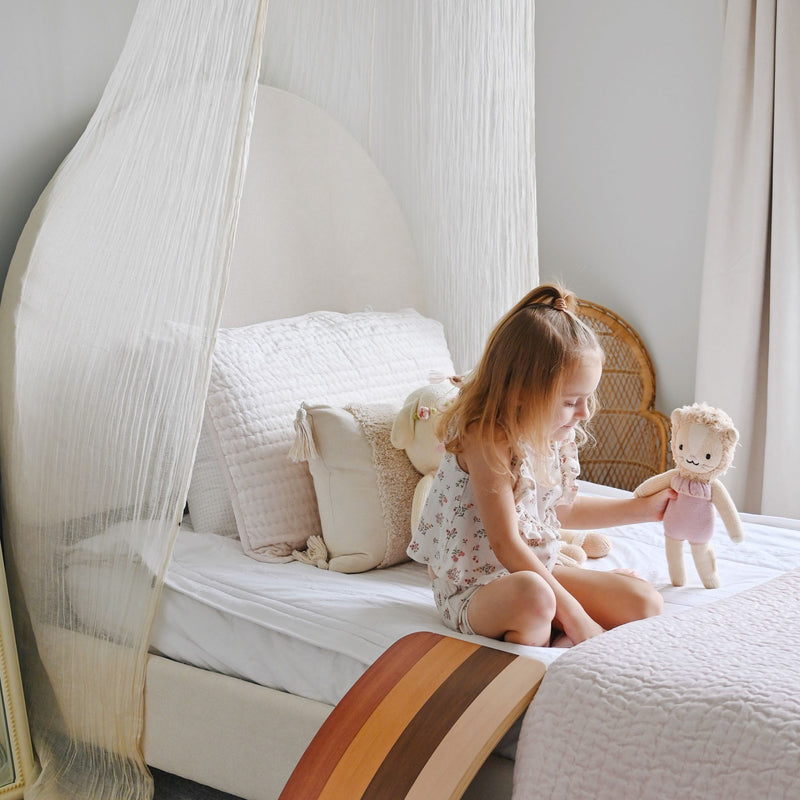 Girl playing with a plush doll on a neutral boho nursery canopy bed with quilted bedding, tassel pillows and a wooden balance board