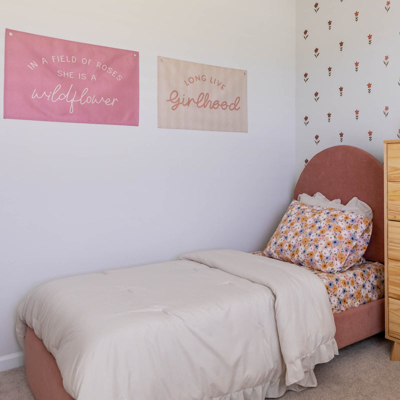 Pink boho girls bedroom with cream comforter, floral sheets and pillow, pink velvet headboard, floral wall decals and inspirational wall banners reading "wildflower" and "girlhood