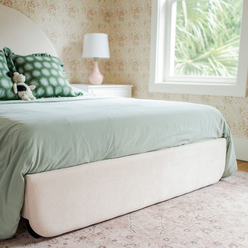 Sage green bedding on an upholstered bed with patterned green pillows, stuffed toy, pink bedside lamp and floral wallpaper by a sunlit window