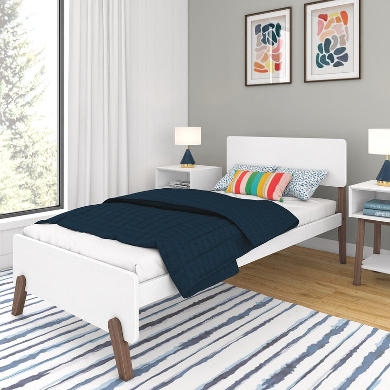 Scandinavian-style kids bedroom with white platform bed, navy quilt, colorful striped pillow, white bedside tables with blue-and-gold lamps, abstract framed wall art, large window with white curtains, hardwood floor and blue-striped area rug