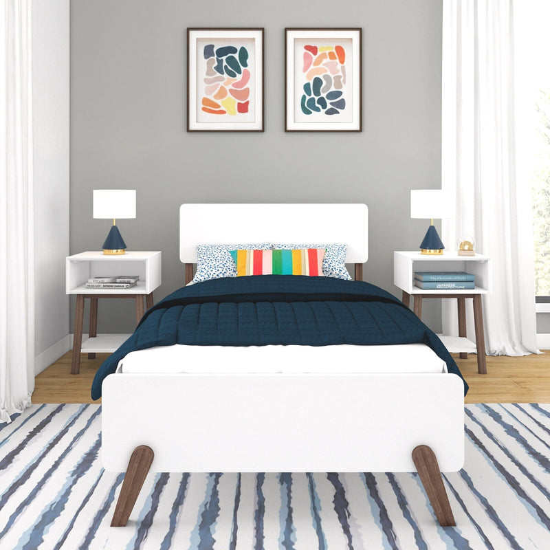 Modern bedroom interior with white platform bed, navy quilt, multicolor striped accent pillow, mid-century nightstands and blue ceramic lamps, abstract framed art on gray wall, and blue-striped area rug