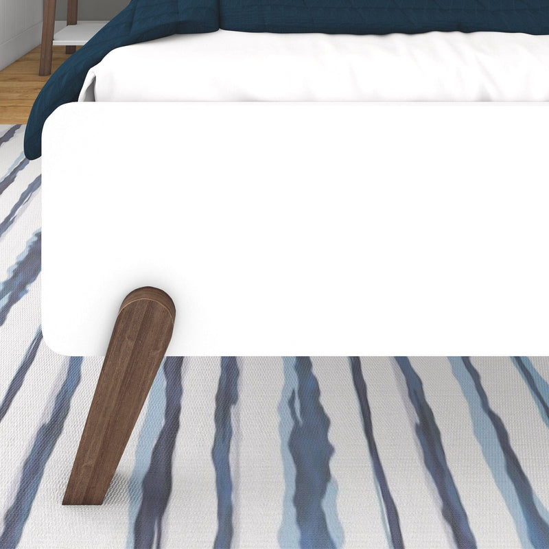 Close-up of white mattress and navy quilt with wooden bed leg on blue-striped area rug, modern bedroom decor