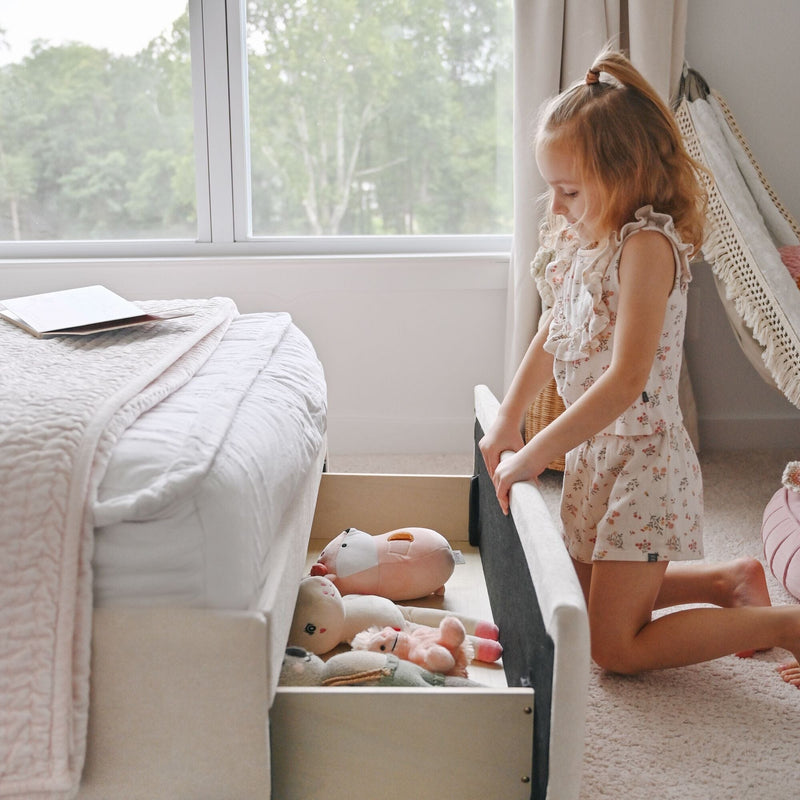 Toddler girl kneeling by a bed in a bright neutral nursery pulling open an under‑bed toy storage drawer filled with plush toys and natural window light