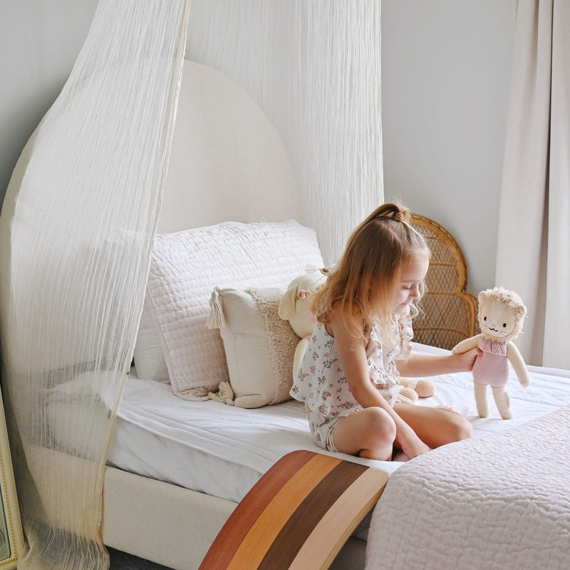 Girl playing with a stuffed doll on a canopy bed in a neutral nursery with quilted bedding, wicker chair and wood rocker
