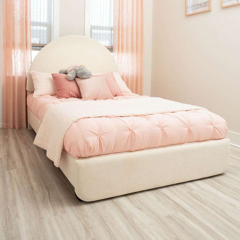 Blush pink bedroom with tufted comforter and velvet pillows, arched upholstered headboard, stuffed hippo toy, sheer peach curtains and light wood floor
