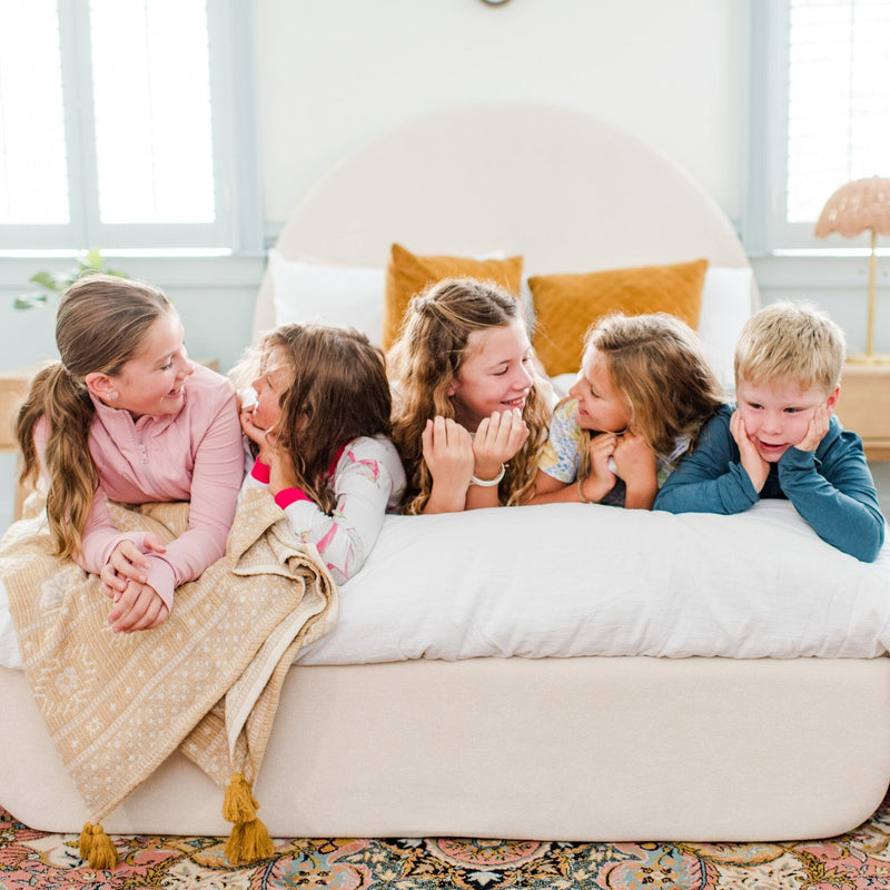 Group of smiling children lying on a cozy bed with pillows and a blanket in a bright bedroom